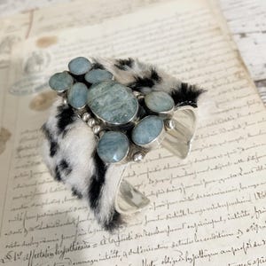May include: A silver cuff bracelet with a black and white faux fur band. The bracelet is adorned with multiple light blue gemstone accents.