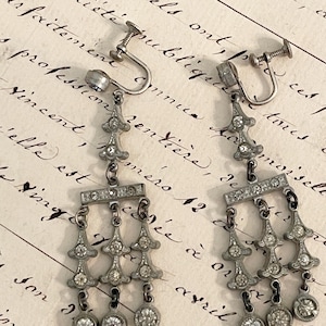May include: A pair of silver-toned metal screw-back earrings featuring a chandelier design. Each earring has a vertical drop with clear, round-cut rhinestones set into the metal frame, ending in three dangling circular rhinestone charms.