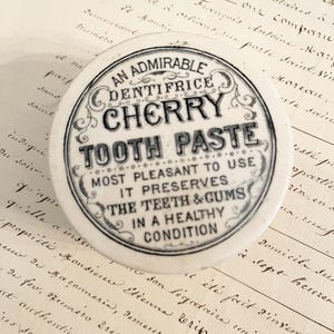 May include: A round, off-white ceramic lid featuring black printed text that reads: AN ADMIRABLE DENTIFRICE CHERRY TOOTH PASTE MOST PLEASANT TO USE IT PRESERVES THE TEETH & GUMS IN A HEALTHY CONDITION, surrounded by decorative scrollwork.