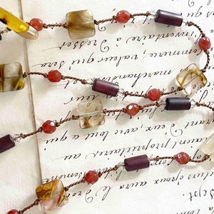 May include: A long, layered necklace with brown cord, square-shaped brown, clear, and white agate beads, and small, dark purple beads. The necklace is on a white surface with brown writing.