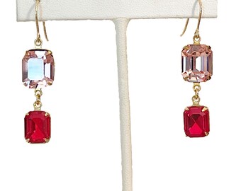 May include: A pair of gold-toned earrings with a two-tiered design. Each earring features a large, rectangular, faceted pink crystal at the top and a smaller, rectangular, faceted red crystal at the bottom. The earrings are displayed on a white stand.