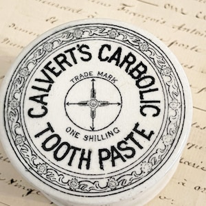 May include: A round white ceramic container lid with black text reading Calvert's Carbolic Tooth Paste and One Shilling. The lid features a central arrow cross symbol and a decorative floral border pattern with a visible fine crackle glaze finish.