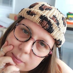 May include: A close-up of a person wearing a brown and black leopard print crochet beanie. The beanie has a wide brim and is made with a chunky yarn. The person is also wearing round, tortoiseshell glasses.