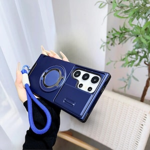 May include: A navy blue phone case with a built-in ring holder and a camera-style design. The case has a matching blue lanyard with a white accent. The phone case has a glossy finish and a raised camera bump.