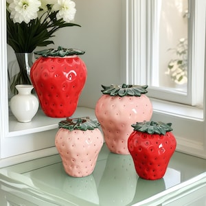 May include: Four ceramic strawberry-shaped containers with green leaves. Two are red and two are pink.
