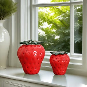 May include: Two red ceramic strawberry-shaped canisters with green leaves on top. The larger canister is on the left and the smaller canister is on the right.