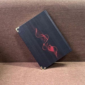 K&ouml;nnte beinhalten: Ein dunkles Buch mit einem roten abstrakten Design von zwei sich ausstreckenden H&auml;nden. Die Ecken des Buches sind mit goldfarbenen Metalleckensch&uuml;tzern verziert. Das Buch liegt auf einer braunen Oberfl&auml;che.