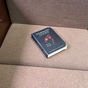 K&ouml;nnte beinhalten: Ein Buch mit dem Titel "DAMAGED GOODS" mit einem dunklen Cover, das zwei Figuren zeigt. Das Buch hat auch den Text "Her scar, His mark" und "SLYTHERIN AFTER DARK". Das Buch liegt auf einer braunen, gepolsterten Oberfl&auml;che.