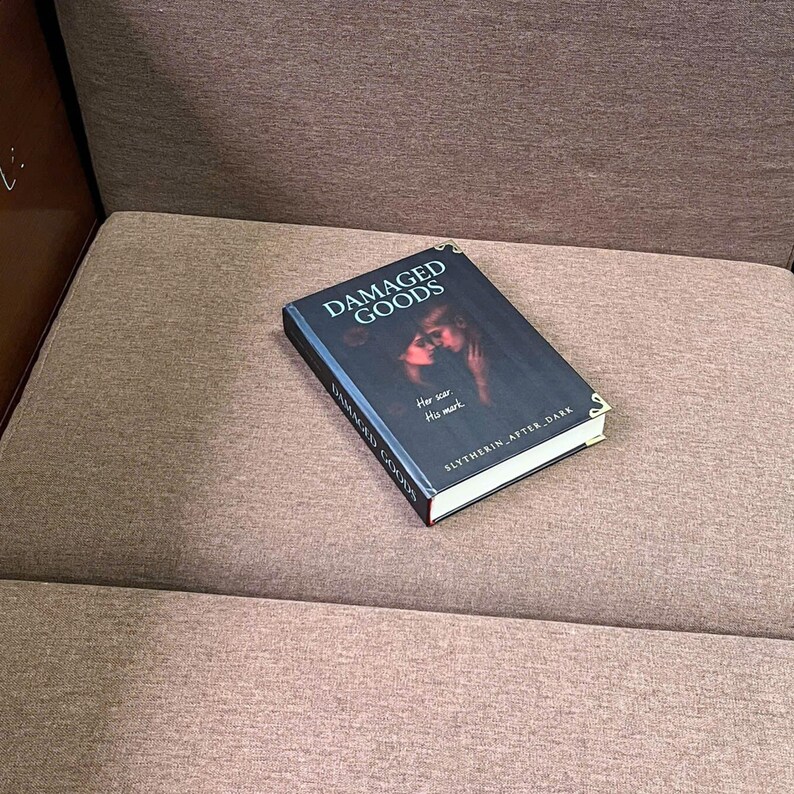 K&ouml;nnte beinhalten: Ein Buch mit dem Titel "DAMAGED GOODS" mit einem dunklen Cover, das zwei Figuren zeigt. Das Buch hat auch den Text "Her scar, His mark" und "SLYTHERIN AFTER DARK". Das Buch liegt auf einer braunen, gepolsterten Oberfl&auml;che.