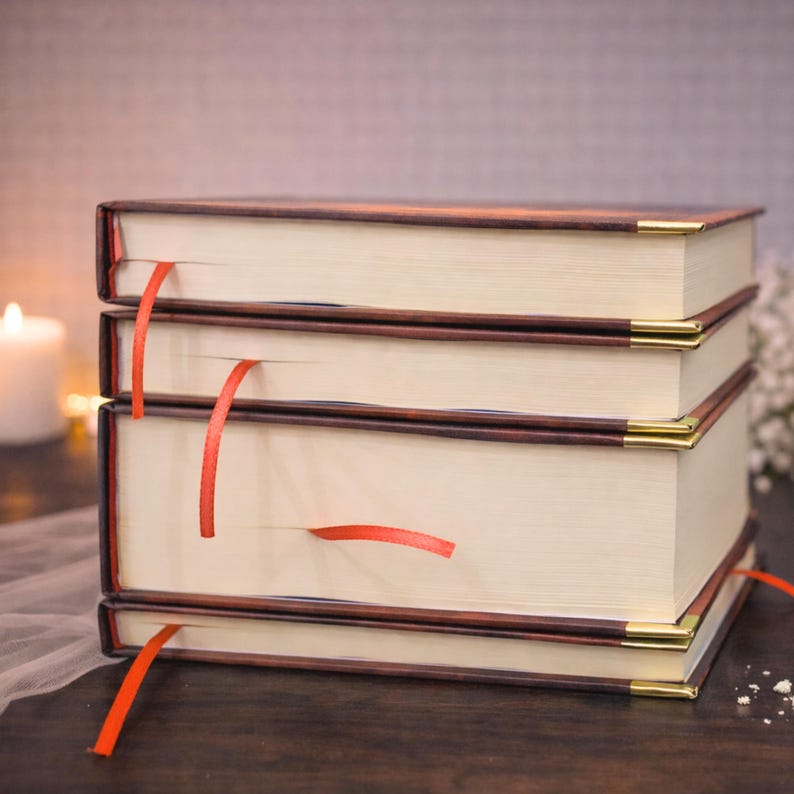 Puede incluir: Una pila de cuatro libros de tapa dura con p&aacute;ginas color crema y cubiertas marrones. Cada libro tiene una cinta roja como marcador. Las esquinas de los libros est&aacute;n acentuadas con metal dorado. Los libros est&aacute;n apilados sobre una superficie de madera oscura.