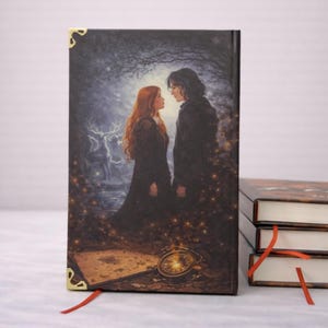 Puede incluir: Un libro de tapa dura con una ilustraci&oacute;n de fantas&iacute;a. La imagen muestra a una pareja en un bosque, con un ciervo y rel&aacute;mpagos en el fondo. El libro tiene esquinas doradas y una cinta roja como marcador. Otros libros est&aacute;n apilados.