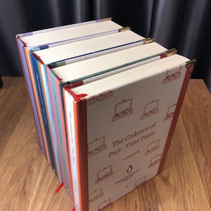 May include: A stack of hardcover books titled "The Cadence of Part-Time Poets" by meawolo. The books have cream-colored covers with a repeating suitcase pattern and gold corner protectors. Each book has a different colored spine and a red ribbon bookmark.