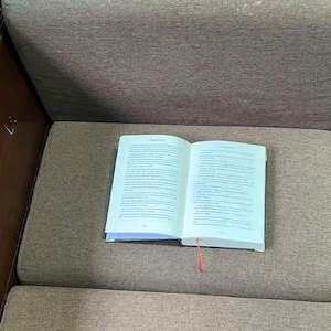 K&ouml;nnte beinhalten: Ein offenes Buch mit Text auf seinen Seiten, das auf einem grauen Sofa liegt. Das Buch hat ein rotes Band als Lesezeichen. Die Seiten sind wei&szlig; mit schwarzem Text. Der Einband des Buches ist dunkel und die Seiten sind leicht vergilbt.
