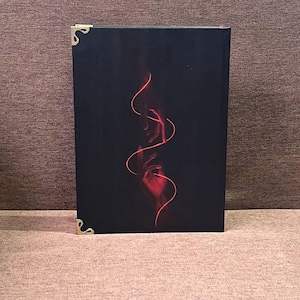 K&ouml;nnte beinhalten: Ein schwarzes Buch mit einem roten abstrakten Design aus H&auml;nden und flie&szlig;enden Linien. Das Buch hat goldene Eckensch&uuml;tzer. Das Cover ist ein auff&auml;lliger Kontrast von Rot vor dunklem Hintergrund.