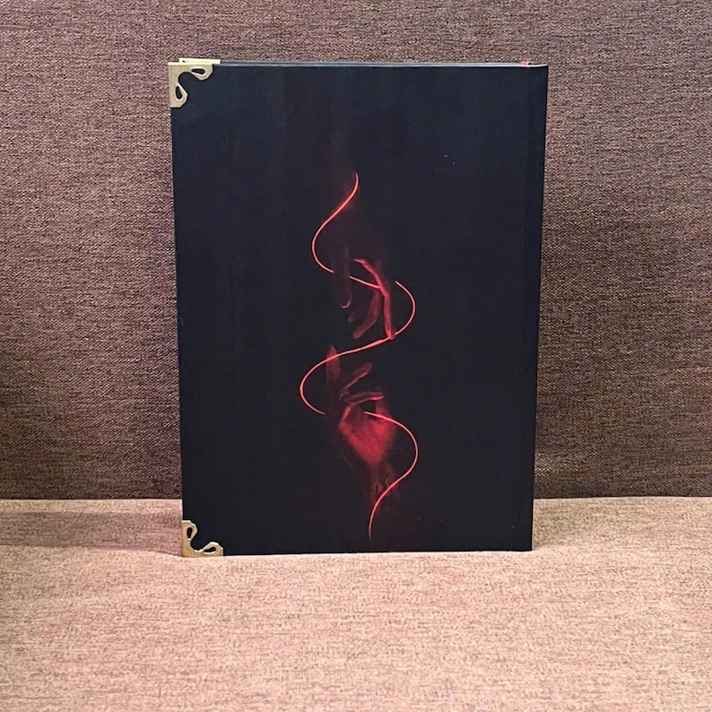 K&ouml;nnte beinhalten: Ein schwarzes Buch mit einem roten abstrakten Design aus H&auml;nden und flie&szlig;enden Linien. Das Buch hat goldene Eckensch&uuml;tzer. Das Cover ist ein auff&auml;lliger Kontrast von Rot vor dunklem Hintergrund.
