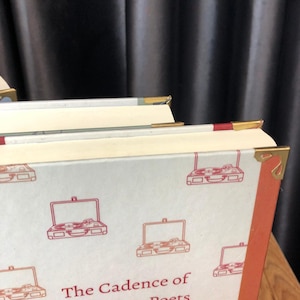 May include: Close-up of a stack of books with white covers featuring a red outline illustration of a record player. The book edges have gold and red accents. The title "The Cadence of Poets" is visible.