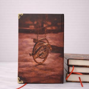 Puede incluir: Una pila de tres libros de tapa dura con marcadores de cinta roja. La portada del libro superior presenta una ilustraci&oacute;n detallada de una esfera armilar dorada colgando de una cadena sobre un fondo marr&oacute;n. Se ven protectores de esquina dorados.