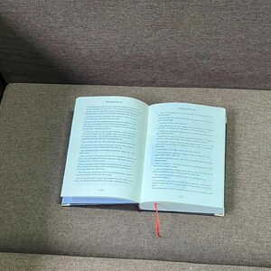 K&ouml;nnte beinhalten: Ein offenes Buch mit Text auf den Seiten und einem roten Leseb&auml;ndchen. Das Buch hat einen schwarzen Einband und liegt auf einer braunen Oberfl&auml;che. Die Seiten sind wei&szlig; mit schwarzem Text.