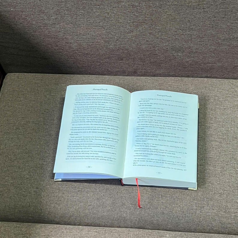 K&ouml;nnte beinhalten: Ein offenes Buch mit Text auf den Seiten und einem roten Leseb&auml;ndchen. Das Buch hat einen schwarzen Einband und liegt auf einer braunen Oberfl&auml;che. Die Seiten sind wei&szlig; mit schwarzem Text.