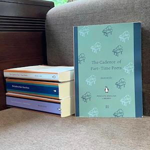 May include: A stack of Penguin English Library books. The front book is titled "The Cadence of Part-Time Poets" with a teal cover and piano illustrations. Other books have orange, blue, and purple spines. The books are on a brown surface.