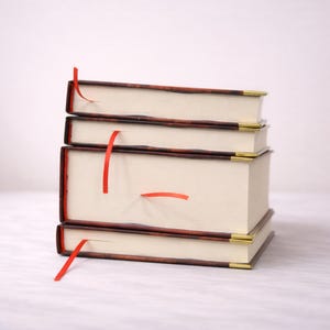 Puede incluir: Una pila de cuatro libros de tapa dura con p&aacute;ginas color crema y cintas rojas como marcadores. Las cubiertas son marrones con un borde marr&oacute;n rojizo y detalles dorados en las esquinas. Los libros est&aacute;n apilados.