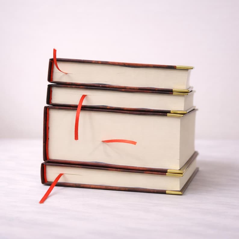Puede incluir: Una pila de cuatro libros de tapa dura con p&aacute;ginas color crema y cintas rojas como marcadores. Las cubiertas son marrones con un borde marr&oacute;n rojizo y detalles dorados en las esquinas. Los libros est&aacute;n apilados.