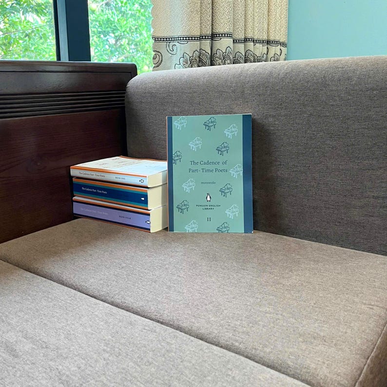 May include: A stack of Penguin English Library books, including "The Cadence of Part-Time Poets" by moranville. The book covers are various colors, with the featured book having a teal background and piano illustrations. The books are on a gray couch.