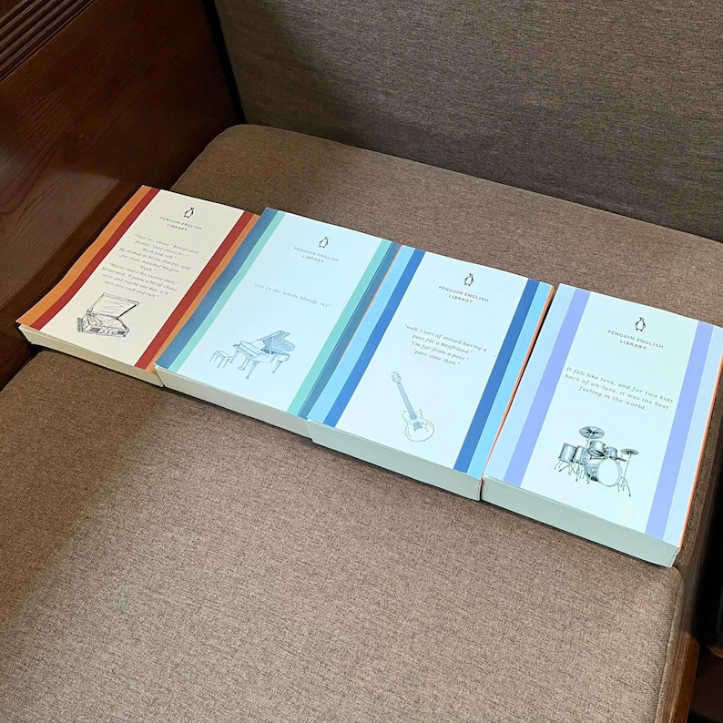 May include: Four Penguin English Library books are displayed, each with a different cover design. The covers feature illustrations of a piano, guitar, and drum set, with text. The books are arranged on a brown surface.