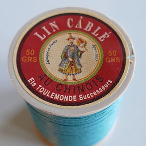 以下が含まれることがあります： 赤とクリーム色のラベルが付いた、ターコイズ色の糸巻き。ラベルには「LIN CABLE AU CHINOIS」と書かれており、カラフルな衣装を着た人物のグラフィックがあります。糸巻きには50グラムの糸が入っており、「Fabriqué en France」と記載されています。