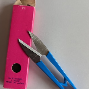 Puede incluir: Un par de tijeras con mangos azules y cuchillas plateadas. Las tijeras están sobre una superficie blanca junto a una caja rosa con el texto "No. 27-07065 1 PIECE MADE IN JAPAN".