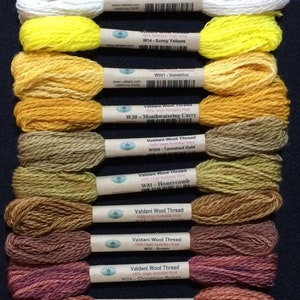 May include: A collection of Valdani Wool Thread skeins in various colors, including ivory, yellow, gold, brown, and cinnamon. Each skein is labeled with its color name and a product code. The threads are arranged vertically against a dark background.