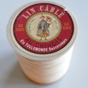 Op de afbeelding: Een spoel crèmekleurig garen met een rood en goud label. Het label leest "LIN CABLE AU CHINOIS" en toont een illustratie van een persoon in traditionele kleding. De spoel bevat 50 gram garen.
