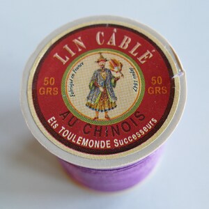 May include: A spool of purple thread with a red and white label. The label reads "LIN CABLE AU CHINOIS" and features an illustration of a man in a colorful outfit. The label also indicates the thread weighs 50 grams.
