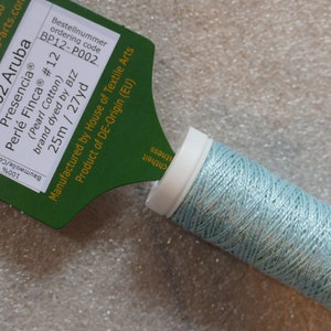 May include: A spool of light blue pearl cotton embroidery thread, labeled "P002 Aruba Presencia® Perlé Finca® #12 (Pearl Cotton) brand dyed by BIZ 25m / 27yd" and "Bestellnummer ordering code BP12-P002".