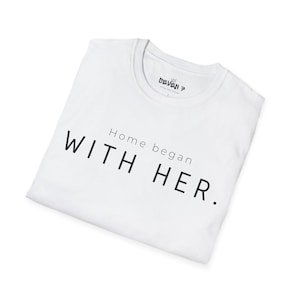 Puede incluir: Una camiseta blanca con el texto "Home began WITH HER." en negro. La camiseta tiene cuello redondo y un pequeño logotipo cerca del cuello. La camiseta está doblada cuidadosamente.