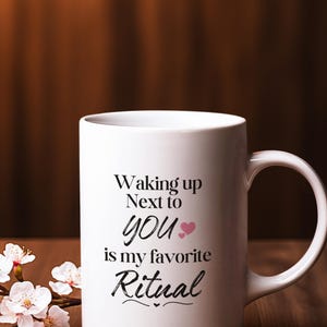 Puede incluir: Una taza de cerámica blanca con el texto "Waking up Next to YOU is my favorite Ritual" en escritura negra. Un corazón rosa está al lado de la palabra "YOU". La taza está sobre una superficie de madera con flores blancas.