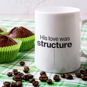Puede incluir: Una taza de café blanca con el texto "His love was structure" en negro, acompañada de tres magdalenas de chocolate en moldes de papel verdes y granos de café esparcidos. Los artículos están colocados sobre un paño a cuadros verde y blanco.
