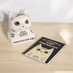 May include: A white ceramic cat figurine with a black outline and text that reads "Phrenology Cat" and lists cat personality traits like "World Domination", "Eat", "Chase Invisible Objects", and "Sleep". A black and white book with the text "The Little Book of Cat Phrenology" is also shown.