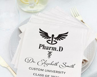 Könnte beinhalten: Weiße Papierservietten mit schwarzem Pharm.D-Logo, dem Namen "Dr. Elizabeth Smith" und dem Aufdruck "Custom University Class of 2026". Die Servietten sind auf einem weißen Teller gestapelt, mit goldenem Besteck.