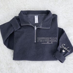 May include: Dark gray quarter-zip sweatshirt with the words "PHYSICIAN assistant" in white block letters on the front. The sleeve cuff has the name "Sophia" and a heart. The sweatshirt is made of a soft material.