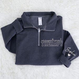 May include: A dark gray quarter-zip sweatshirt with the words "occupational THERAPY" in white block letters. The sleeve cuff has the name "Sophia" in script with a small white heart.