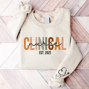 May include: A beige sweatshirt with the text "CLINICAL nurse EST. 2025" in colorful letters. The sweatshirt has a small stethoscope and heart graphic on the sleeve.