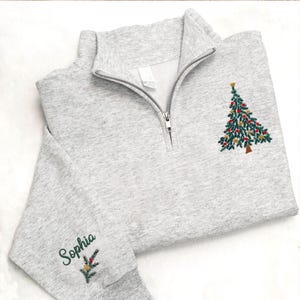 May include: A light gray quarter-zip sweatshirt with a Christmas tree embroidered on the front and the name "Sophia" embroidered on the sleeve. The Christmas tree is green with red and gold ornaments and a gold star on top.