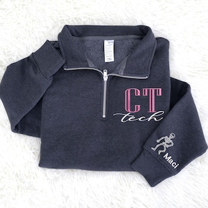 May include: Dark gray pullover sweatshirt with a quarter-zip neckline. The front features the text "CT tech" in pink and white script. The right sleeve has a skeleton graphic and the name "Maci" in white. The sweatshirt is laid flat on a white, fluffy surface.
