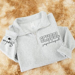 May include: A light gray sweatshirt with a quarter-zip neckline. The text "SCHOOL psychologist" is printed in black on the front. The sleeve cuff has the name "Sophia" and a heart printed in black. The sweatshirt is lying on a brown, textured surface.