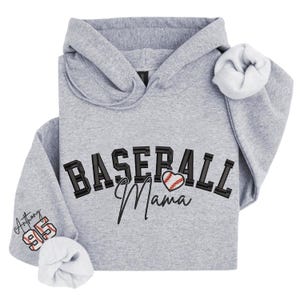 May include: A gray hoodie with the words "BASEBALL Mama" in black and a baseball heart graphic. The sleeve features the name "Anthony" and the number "95" in red and white. The hoodie has a hood and white lining.