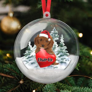 May include: Clear Christmas ornament with a red ribbon. Inside, a brown puppy wearing a Santa hat holds a red heart with the name "Charlie". Snowy trees are in the background.
