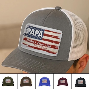 May include: A gray and white trucker hat with a rectangular patch featuring a distressed American flag design. The word "PAPA" is above the flag, with the names Sophia, Emma, John, and Ethan below. The hat has a curved brim.