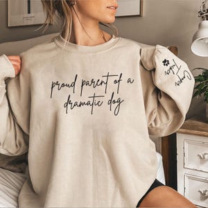 May include: Beige sweatshirt with the phrase "proud parent of a dramatic dog" in black script. The sleeve features a paw print and the names "Tucker" and "Cooper". A casual, comfortable top for dog lovers.