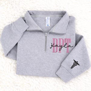 May include: A light gray sweatshirt with a quarter-zip neckline. The front features the embroidered letters "DPT" in pink, with the name "Kayla" in a cursive font underneath. The sleeve has a black caduceus symbol.